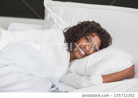 African American woman smiling and reclining on made bed in bedroom under blanket with pillow 135712382