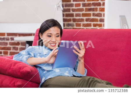 Chinese woman reclining on red upholstered sofa in living room tapping tablet under lamp by monitor Chinese woman reclining on red upholstered sofa in living room tapping tablet under lamp by monitor 135712386