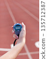 Translucent blue digital stopwatch with black lanyard ticking in hand above red running track lanes 135712387