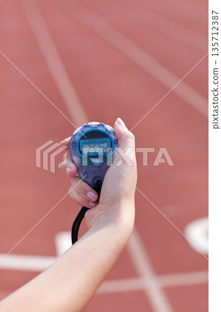 Translucent blue digital stopwatch with black lanyard ticking in hand above red running track lanes Translucent blue digital stopwatch with black lanyard ticking in hand above red running track lanes 135712387