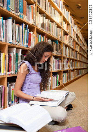 Female student sitting on carpeted library floor studying open textbooks against wooden bookshelves Female student sitting on carpeted library floor studying open textbooks against wooden bookshelves 135712391