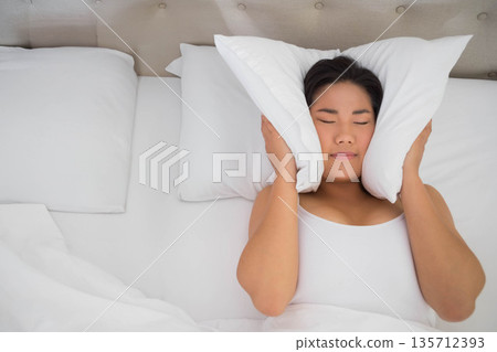Asian woman reclining on bed pressing two white pillows against head with white bedding, headboard 135712393