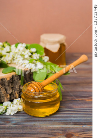 Sweet honey jar surrounded spring acacia blossoms. Honey flows from a spoon in a jar. jars of clear fresh acacia honey on wooden background Sweet honey jar surrounded spring acacia blossoms. Honey flows from a spoon in a jar. jars of clear fresh acacia honey on wooden background 135712449
