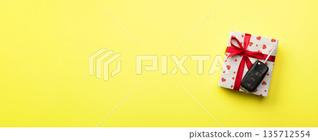 Give gift car key concept top view. Present box with red ribbon bow, heart and car key on yellow colored background Give gift car key concept top view. Present box with red ribbon bow, heart and car key on yellow colored background 135712554