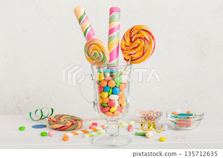 Colorful candies in cup on table on light background background. Large swirled lollipops. Creative concept of a jar full of delicious sweets from the candy store Colorful candies in cup on table on light background background. Large swirled lollipops. Creative concept of a jar full of delicious sweets from the candy store 135712635