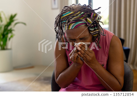 African American senior woman sneezing into white tissue while sitting at home near potted plant 135712854