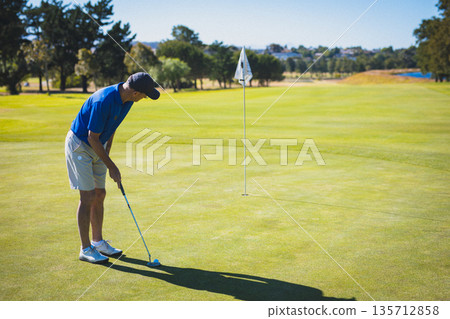 Senior golfer lining up putt on putting green with putter, golf ball near flagstick, copy space 135712858