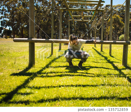 Woman navigating horizontal beam obstacle on wooden posts on grassy field wearing military uniform 135712859
