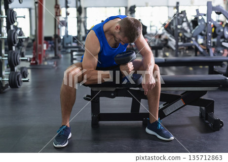 Man sitting on weight bench performing concentration curls in gym wearing sportswear with dumbbell Man sitting on weight bench performing concentration curls in gym wearing sportswear with dumbbell 135712863
