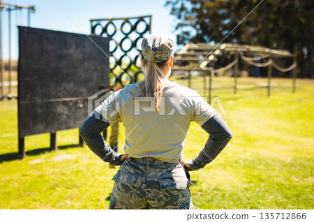 Female service member wearing camouflage uniform standing on field surveying obstacle course in sun Female service member wearing camouflage uniform standing on field surveying obstacle course in sun 135712866