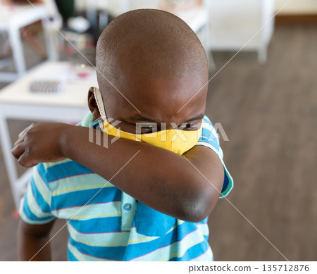 African American boy coughing into elbow, wearing yellow face mask in classroom with white tables 135712876