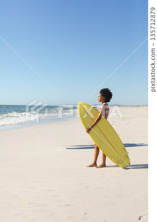 African American woman wearing bikini holding yellow surfboard facing waves on beach, copy space 135712879