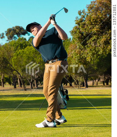Adult male golfer swinging driver and wearing golf glove on fairway by bag under blue sky Adult male golfer swinging driver and wearing golf glove on fairway by bag under blue sky 135712881