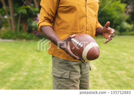 Senior African American man wearing mustard shirt holding brown American football in garden 135712883
