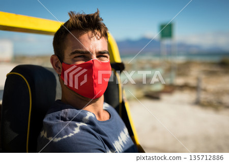 Man sitting on seat frame under yellow roll bar on coastal road wearing red cloth mask 135712886