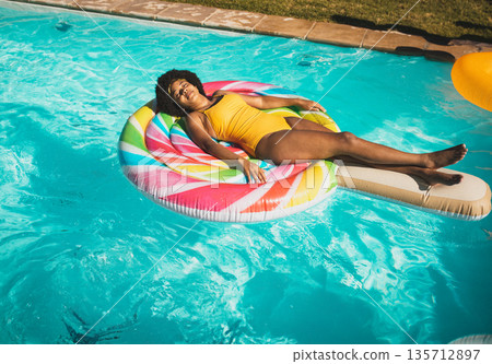African American woman wearing yellow swimsuit relaxing on rainbow float in pool near stone coping African American woman wearing yellow swimsuit relaxing on rainbow float in pool near stone coping 135712897