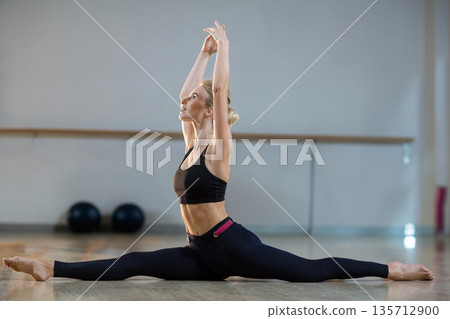 Female dancer performing split stretch on wooden floor in studio with barre and exercise balls 135712900