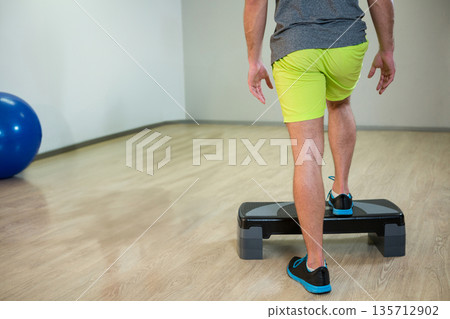 Man in neon shorts stepping onto platform in fitness studio with stability ball, copy space Man in neon shorts stepping onto platform in fitness studio with stability ball, copy space 135712902