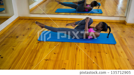 African American woman in gym activewear lying on mat pulling knee toward chest, mirror, copy space African American woman in gym activewear lying on mat pulling knee toward chest, mirror, copy space 135712905