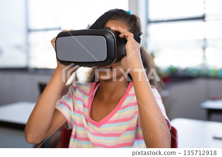 Girl in pastel shirt adjusting virtual reality headset and sitting at desk near blinds and chairs Girl in pastel shirt adjusting virtual reality headset and sitting at desk near blinds and chairs 135712918