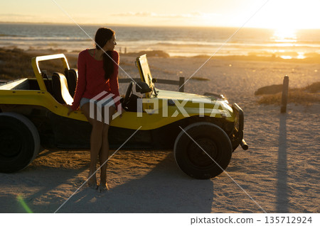 Woman sitting on yellow dune buggy at beach at sunset, gazing ocean and posts, copy space 135712924