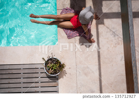 Woman sitting beside rectangular pool on sarong on patio wearing sun hat near planter and bench 135712927