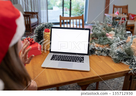 Teen girl wearing Santa hat raising hand greeting laptop at dining table with wreaths, copy space 135712930
