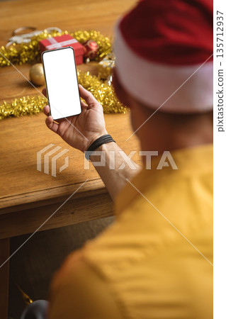 Man sitting at home dining table holding smartphone above tinsel, gift box with baubles, copy space 135712937