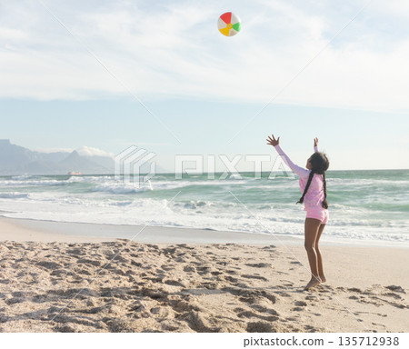 African American girl standing on sandy beach reaching for colorful beach ball overhead, copy space 135712938