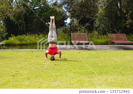 Adult male balancing headstand on lawn, wearing red tee beside wooden deck chairs overlooking pond Adult male balancing headstand on lawn, wearing red tee beside wooden deck chairs overlooking pond 135712942