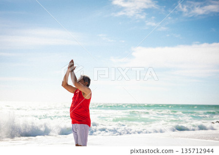 Senior man in red shirt stretching arms overhead at sandy shoreline with ocean waves, copy space Senior man in red shirt stretching arms overhead at sandy shoreline with ocean waves, copy space 135712944