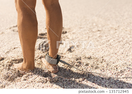 Man standing barefoot on coarse beach sand near shoreline, wearing neoprene surfboard leash cuff 135712945