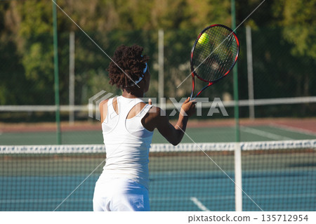 African American woman tennis player swinging racket hitting yellow ball behind net on court 135712954