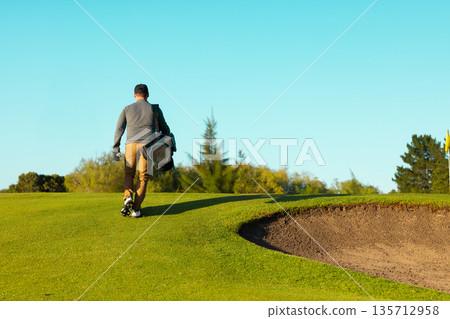 Adult male golfer walking on course with baseball cap, golf shoes, golf bag toward flagstick bunker Adult male golfer walking on course with baseball cap, golf shoes, golf bag toward flagstick bunker 135712958