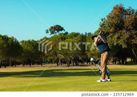 Male golfer swinging club following through on fairway at golf course with golf bag, copy space Male golfer swinging club following through on fairway at golf course with golf bag, copy space 135712964