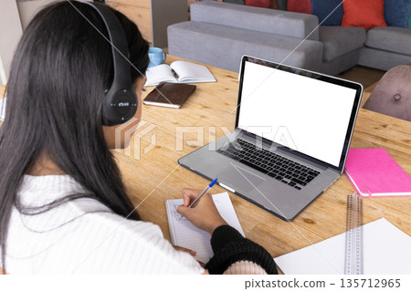 Teenage girl wearing white sweater black headphones writing in notebook at home study with laptop Teenage girl wearing white sweater black headphones writing in notebook at home study with laptop 135712965