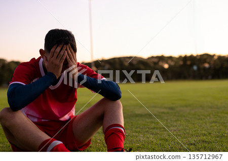 Teenage male soccer player sitting on grass field at dusk shielding face near floodlight poles Teenage male soccer player sitting on grass field at dusk shielding face near floodlight poles 135712967