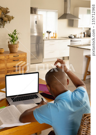 African American teenage boy sitting at table in kitchen studying with laptop, textbooks and papers 135712968