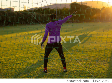 Teenage male goalkeeper wearing padded gloves, pointing on grass pitch by soccer net at golden hour 135712969