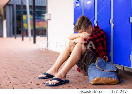 School boy sitting on walkway at school leaning against blue lockers with backpack, copy space School boy sitting on walkway at school leaning against blue lockers with backpack, copy space 135712974