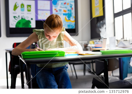 School-aged girl leaning forward at desk in classroom, writing in notebook with green pencil case School-aged girl leaning forward at desk in classroom, writing in notebook with green pencil case 135712976