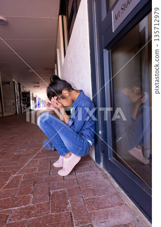 Asian child crouching and covering face on brick corridor floor beside Art Studio door, copy space 135712979