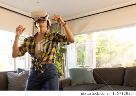 Woman using virtual reality headset interacting with environment in living room near sectional sofa Woman using virtual reality headset interacting with environment in living room near sectional sofa 135712983
