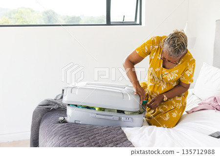 Senior African American woman kneeling packing suitcase on bed with smartphone and lamp, copy space Senior African American woman kneeling packing suitcase on bed with smartphone and lamp, copy space 135712988