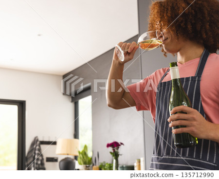 African American adult woman in apron holding wine bottle sipping from glass in kitchen, copy space 135712990