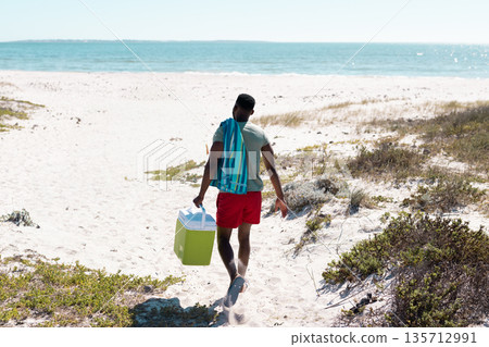 African American man walking across dune toward sea in trunks carrying green cooler, striped towel African American man walking across dune toward sea in trunks carrying green cooler, striped towel 135712991