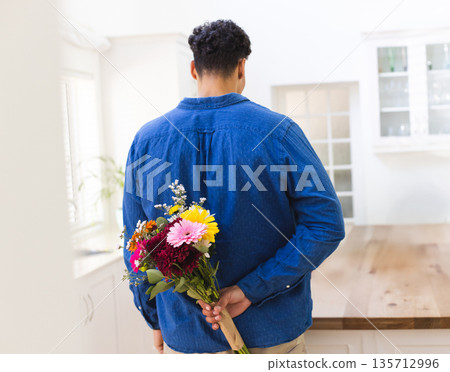 Man in denim and khakis standing in kitchen holding kraft paper wrapped bouquet behind back 135712996