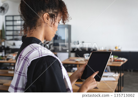 Mid adult woman tapping tablet while draping tea towel by wooden tables in restaurant, copy space 135713002