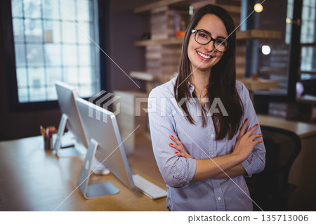 Smiling woman wearing shirt standing arms crossed by desk at office with monitors and pencils 135713006