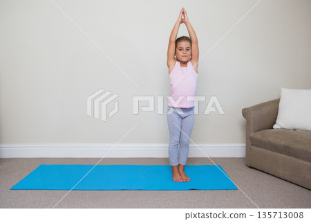 Female child performing full-body stretch on blue yoga mat at home with brown couch, white pillow 135713008